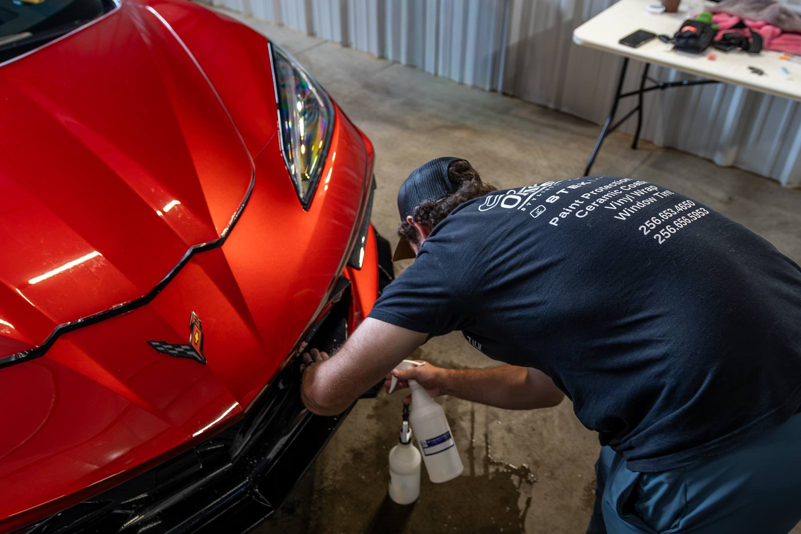 Detailer working along the front of a red Corvette inside the shop, spray bottles at hand